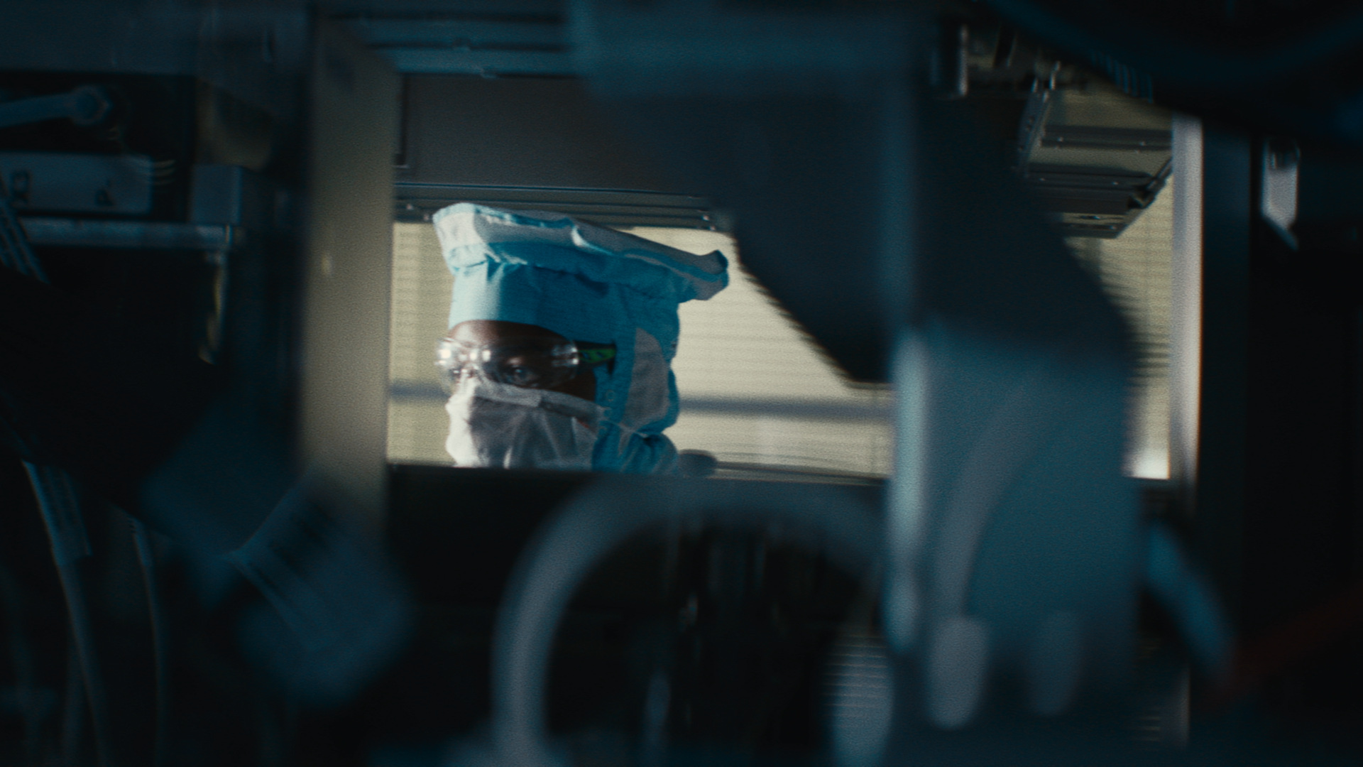 The camera looks through a machine to see the face of a man wearing a cleanroom suit, a mask and safety goggles. The camera looks through a machine to see the face of a man wearing a cleanroom suit, a mask and safety goggles.