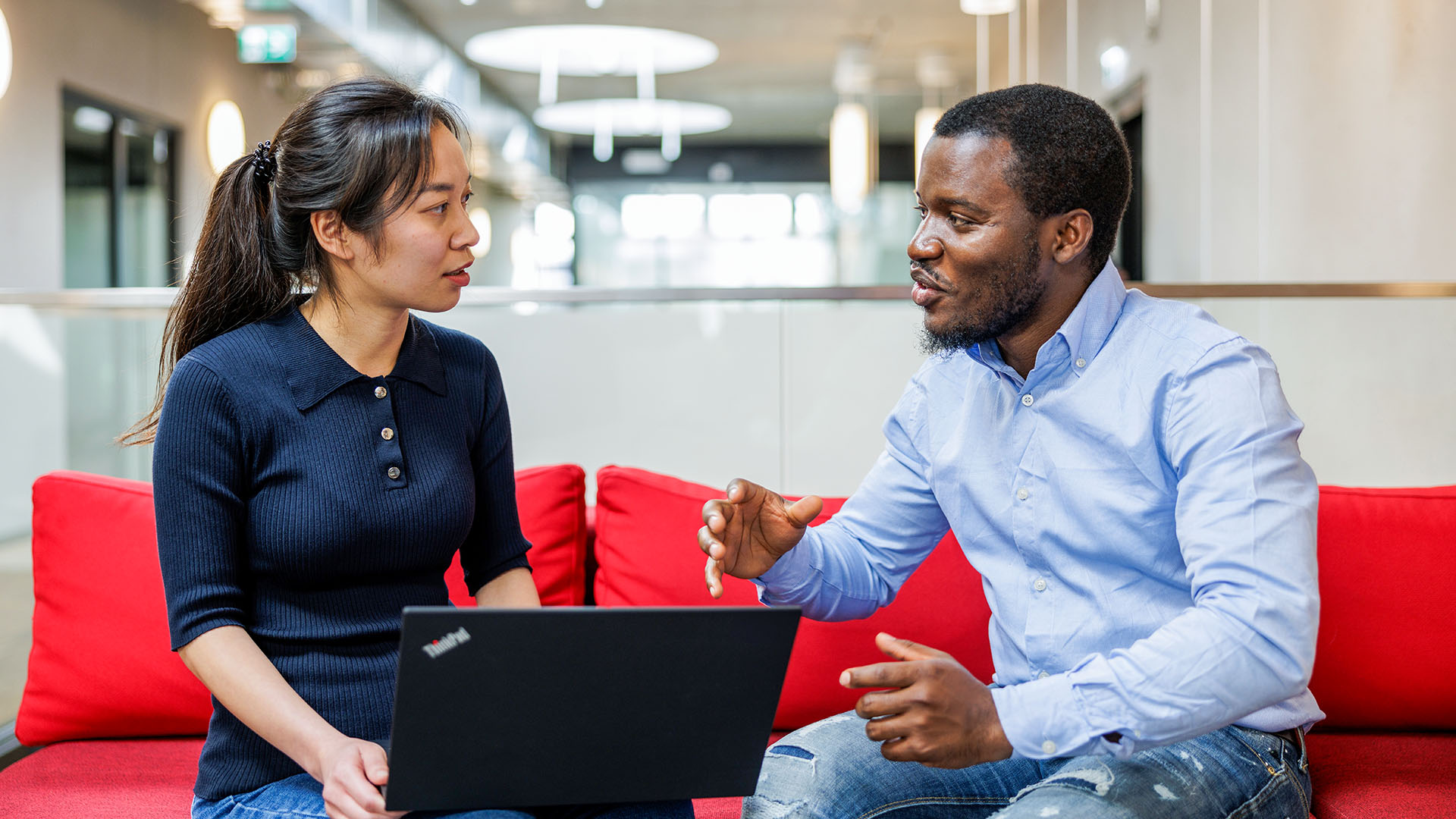 Two ASML employees engaging in a discussion while sitting on a red couch in a modern office space, with one person holding a laptop.