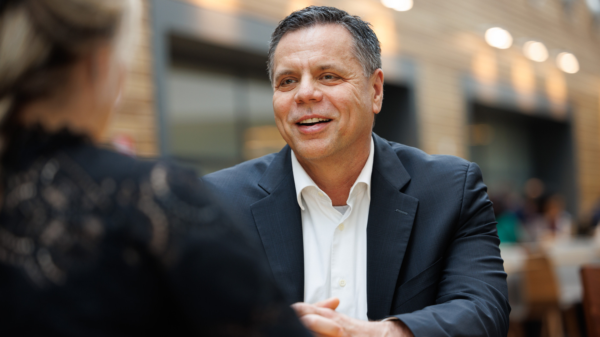A man wearing a dark blue suit and white shirt with no tie sits at a table in a cafeteria and smiles at his interviewer. A man wearing a dark blue suit and white shirt with no tie sits at a table in a cafeteria and smiles at his interviewer.