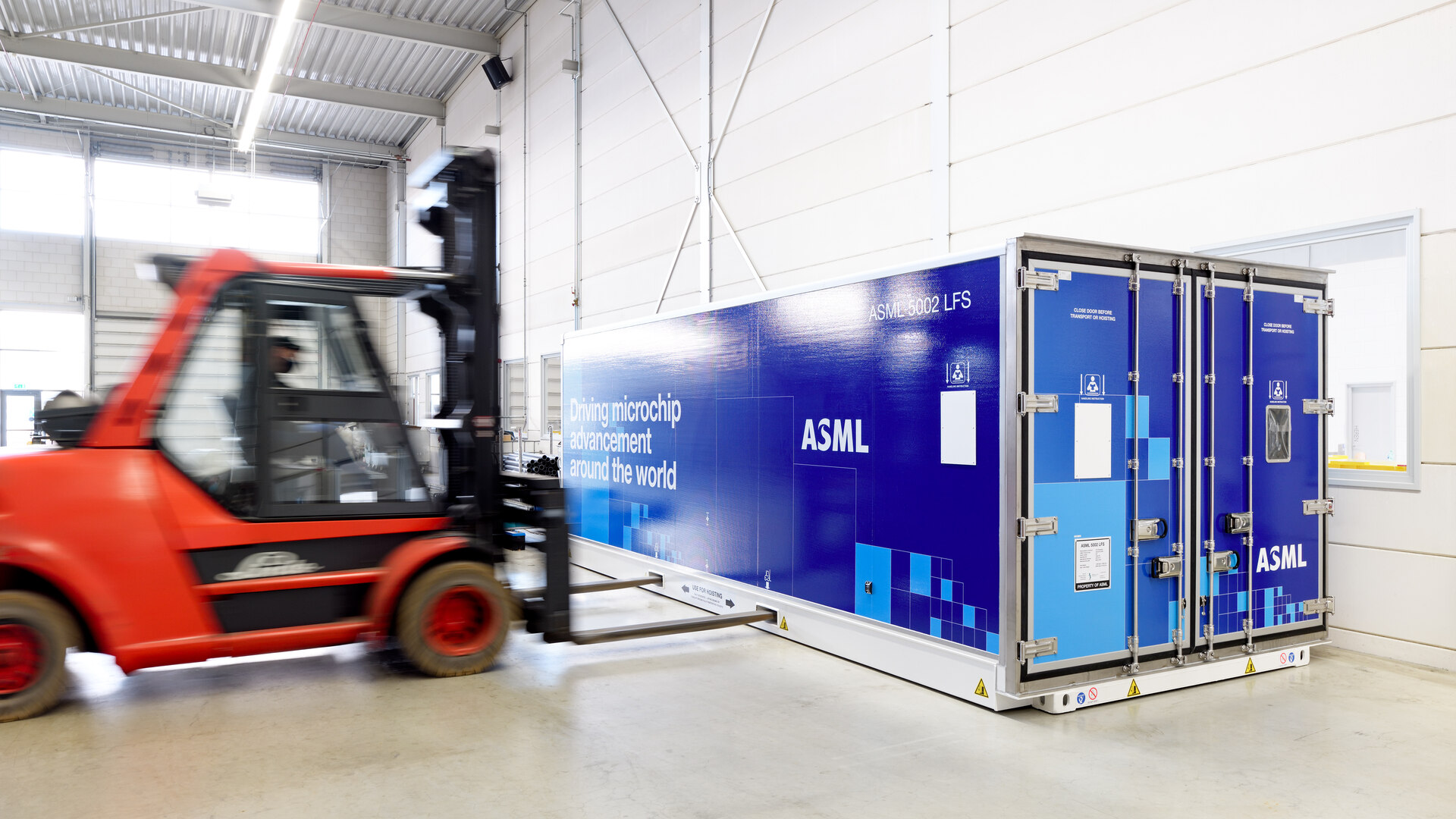 A forklift inside a warehouse approaches a large blue container bearing the words “ASML” and “Driving microchip advancement around the world”. A forklift inside a warehouse approaches a large blue container bearing the words “ASML” and “Driving microchip advancement around the world”.