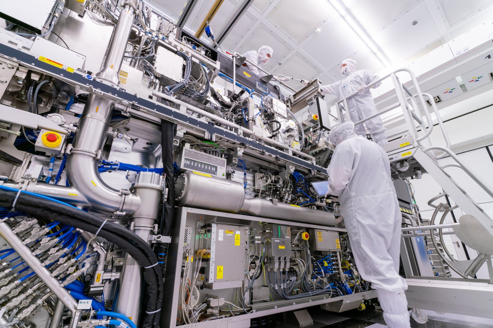 a low-angle photo of three people in cleanroom suits working on a huge lithography system with many wires and pipes. a low-angle photo of three people in cleanroom suits working on a huge lithography system with many wires and pipes.