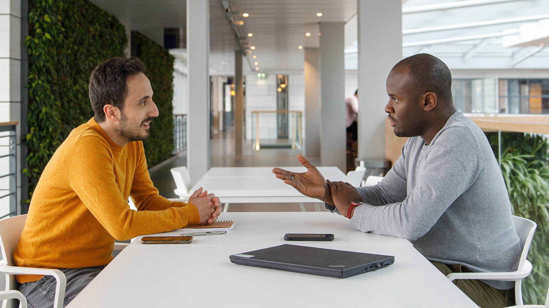 Two ASML colleagues engaging in a conversation at a white table, with one person explaining something with his hands, and a closed laptop, suggesting a collaborative work session. 