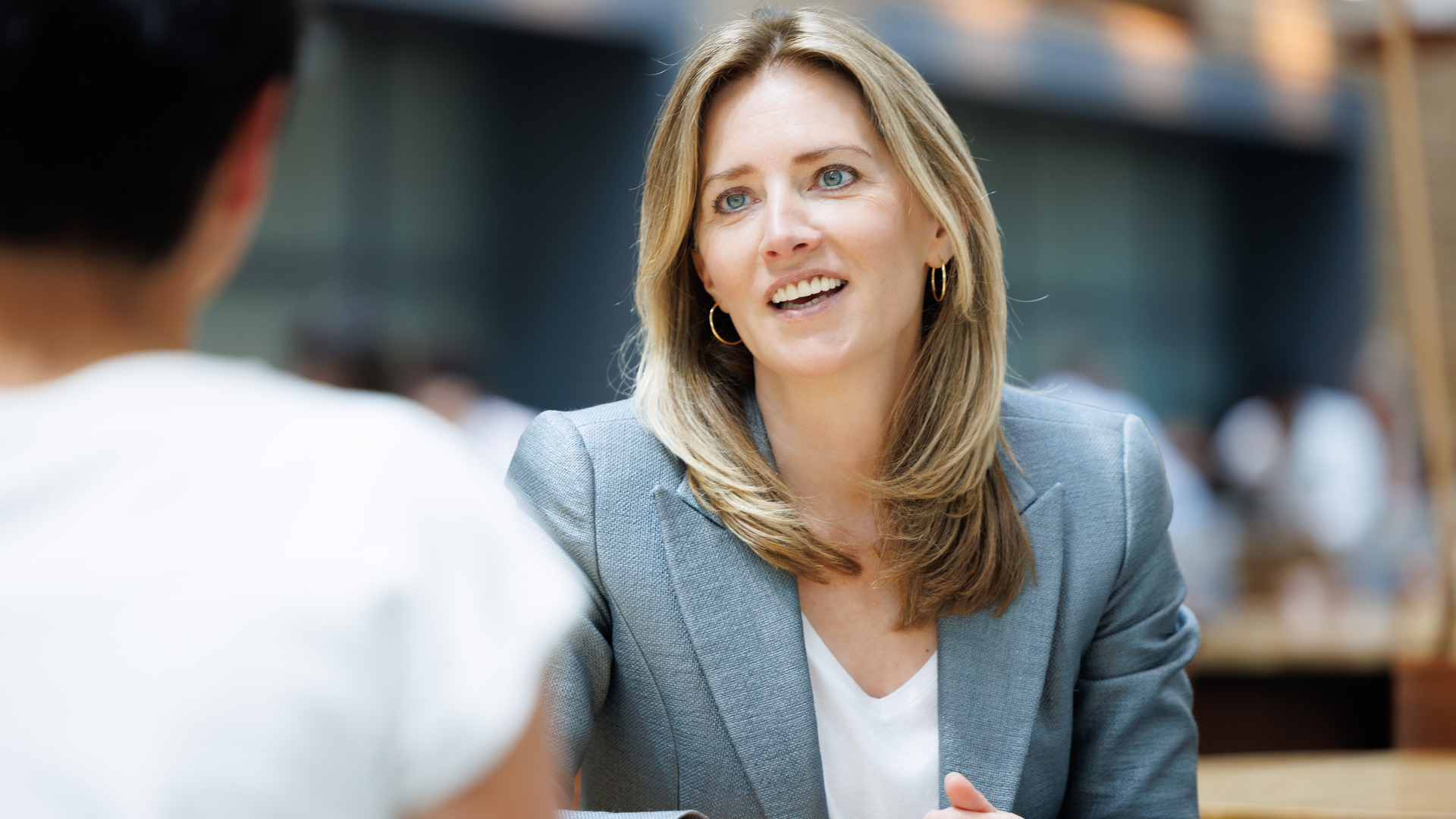 A blonde woman in a gray blazer and white shirt sits at a table in a cafeteria and smiles at her interviewer. A blonde woman in a gray blazer and white shirt sits at a table in a cafeteria and smiles at her interviewer.
