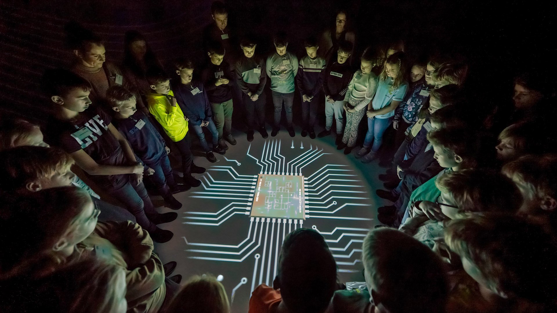 A group of children and adults standing around a projection of a microchip A group of children and adults standing around a projection of a microchip