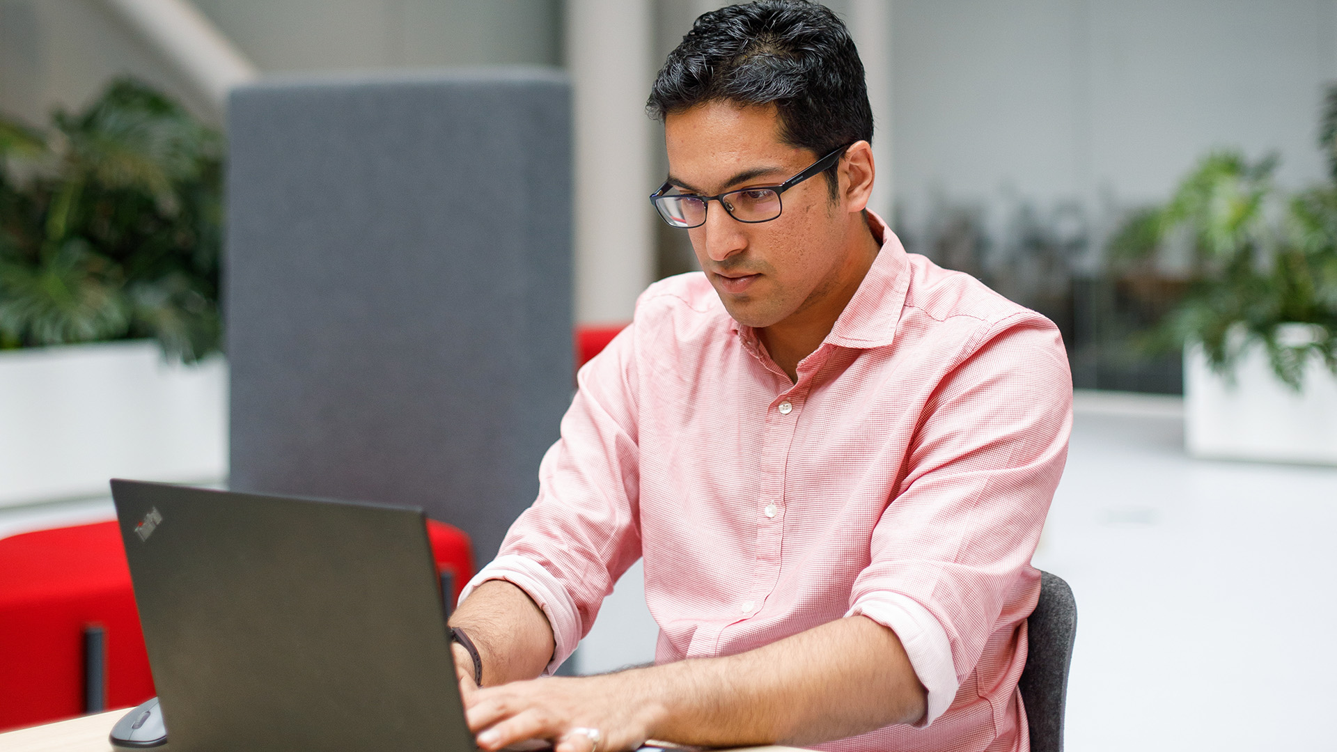An ASML employee wearing glasses and a pink shirt sitting at a desk and working on a laptop.