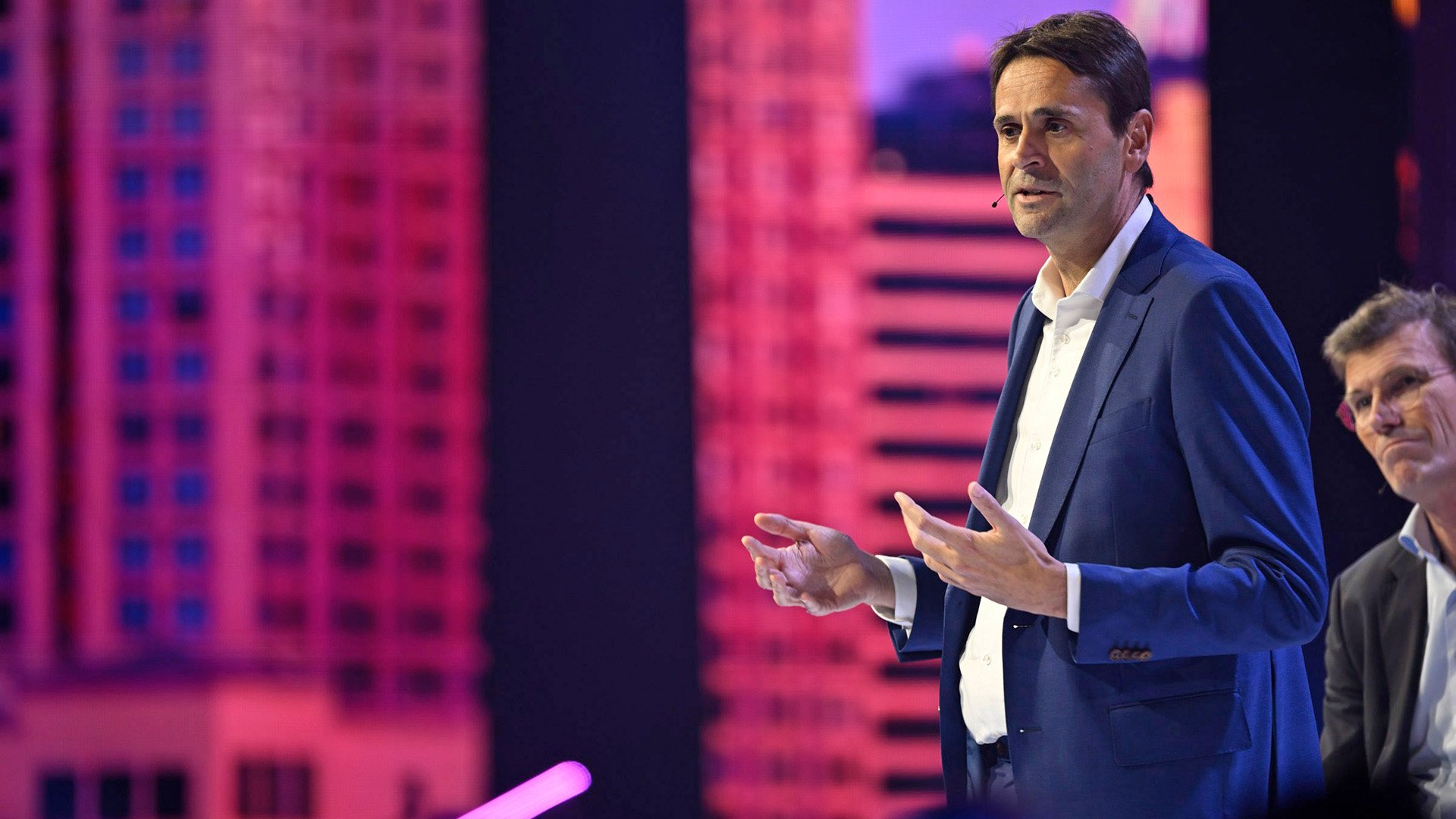 Christophe Fouquet, dressed in a navy blue suit and white shirt, gesturing with open hands against a vibrant pink digital backdrop, communicating a point during a presentation.