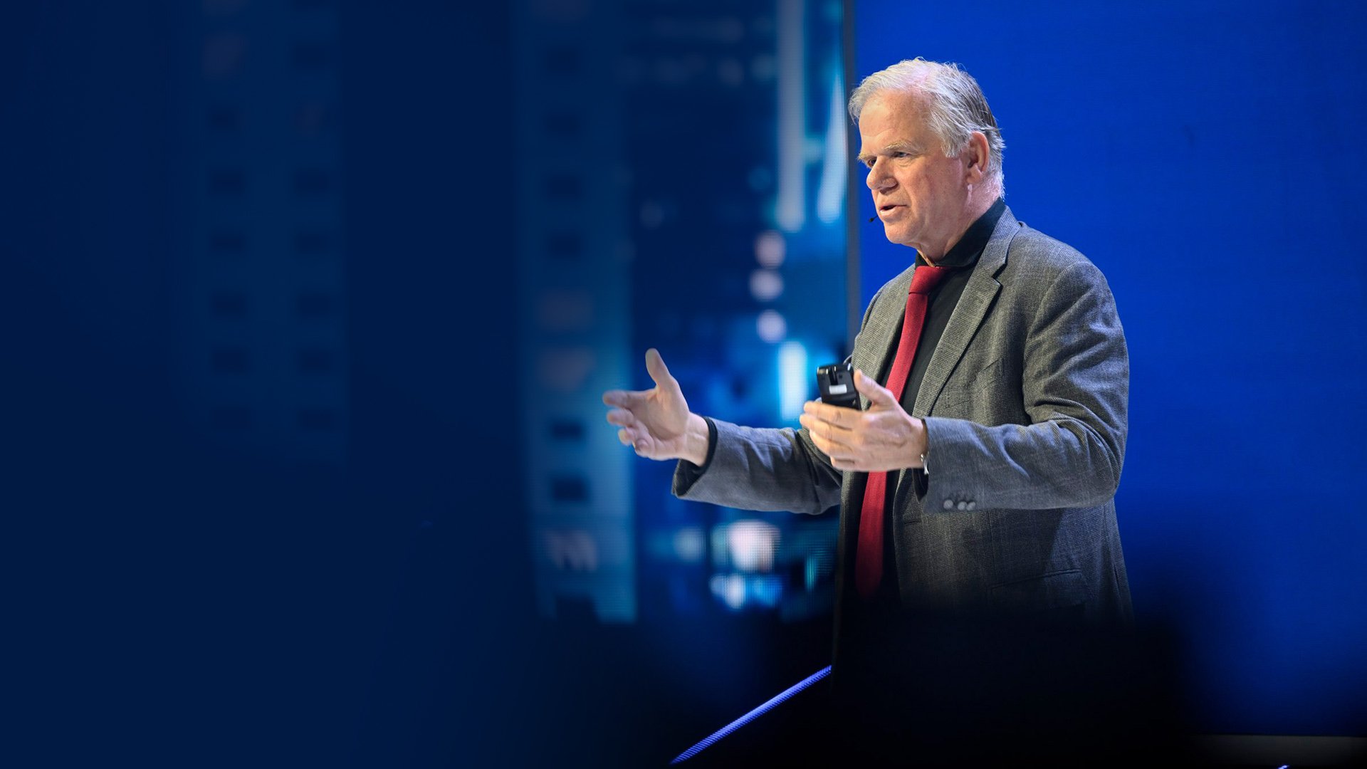 Martin van der Brink giving a presentation with a remote in hand, dressed in a grey jacket and red tie.