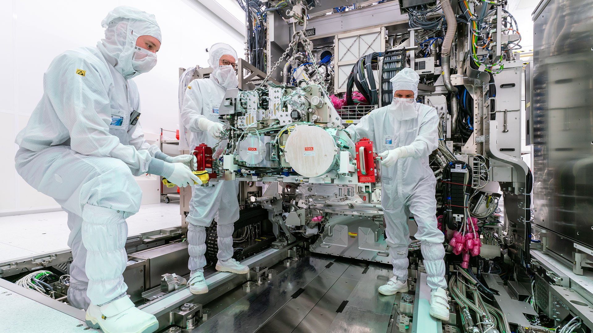 Three cleanroom engineers work on assembling an EUV system, moving the source into the body. Three cleanroom engineers work on assembling an EUV system, moving the source into the body.