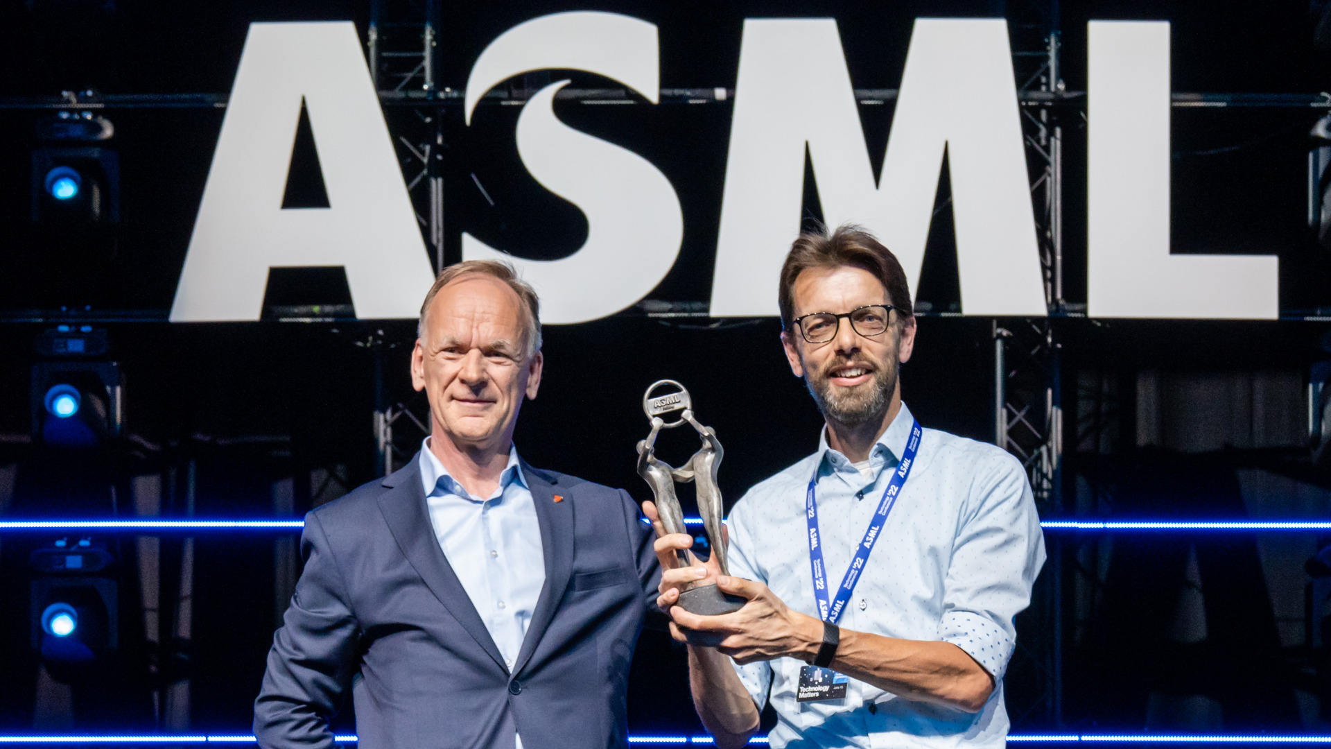ASML Fellow Antoine Kempen stands to the right of senior vice president for technology Jos Benschop. He holds up his fellow award. They both stand under the ASML logo. ASML Fellow Antoine Kempen stands to the right of senior vice president for technology Jos Benschop. He holds up his fellow award. They both stand under the ASML logo.