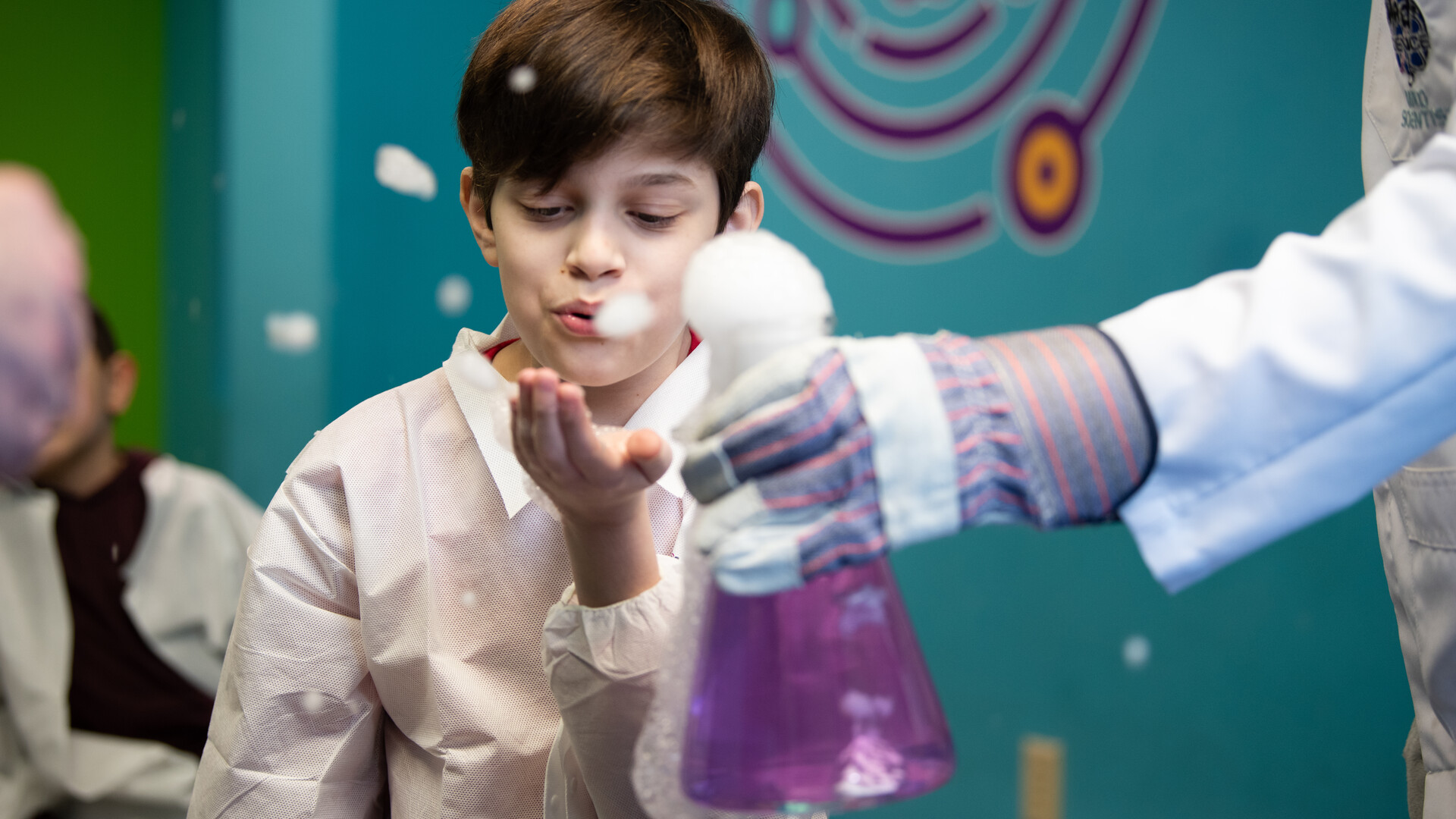 A child is seen blowing at the foam in his hand as a result of an overflowing flask full of bubbly purple liquid during a science experiment. A child is seen blowing at the foam in his hand as a result of an overflowing flask full of bubbly purple liquid during a science experiment.
